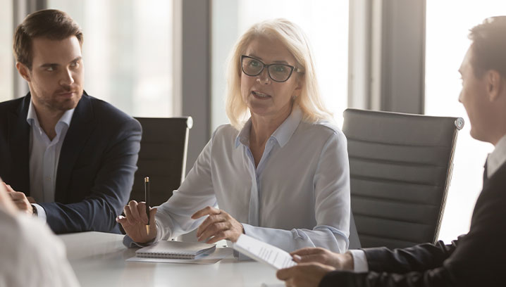 Frau spricht im Meeting, strahlt Autorität aus, Männer hören aufmerksam zu. Spannung und Konzentration im Raum.