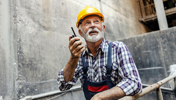 Ein älterer Bauarbeiter mit einem Funkgerät, mit einem gelben Helm in einer Baustelle.