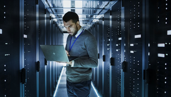 Ein Mann in einem Rechenzentrum arbeitet konzentriert an seinem Laptop zwischen Serverregalen.